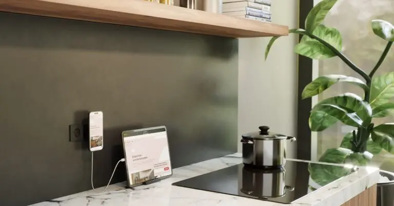 A wireless Qi2 charger plugged into a wall above a kitchen countertop, with a phone attached to it and a tablet charging via wired connection.